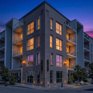 Modern multi-story building w/ private balconies & lush greenery out front.