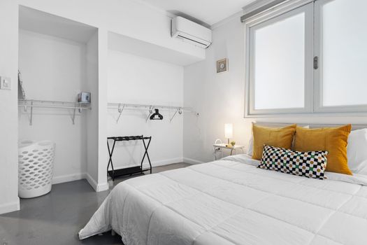 Dormitorio elegante y minimalista con un toque de color, que cuenta con una cómoda cama con almohadas estampadas en color mostaza, complementada con un práctico armario abierto y una moderna unidad de aire acondicionado para una comodidad óptima.