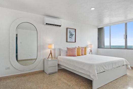 Bedroom with queen bed, white bedding, nightstand, modern lamp, and large oval mirror. Air conditioning unit visible.