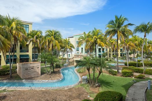 Escape to our tropical oasis with lush gardens and a meandering pool, framed by vibrant buildings and palm trees.