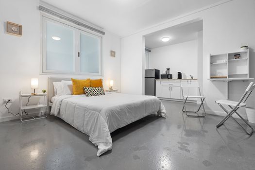 Acogedora vista de entrada de un moderno apartamento tipo estudio, que muestra una cama bien equipada con ropa de cama elegante, iluminación convenientemente ubicada y un vistazo al espacio simplista de la cocina.