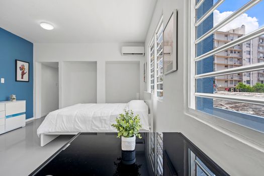 The immaculately clean and modern bedroom features white walls complemented by one blue accent wall. A neatly made white bed is centrally positioned against the accent wall. Above the bed, there is an air conditioning unit installed for comfort. To the left of the bed is a white cabinet adorned with decorative items. A piece of modern artwork hangs on the blue wall adding an artistic touch to the room. A large window allows ample natural light to flood into space, enhancing its brightness. Outside the window are views of buildings indicating an urban setting. On a glossy black surface near the window sits a potted green plant adding a touch of nature to space.
