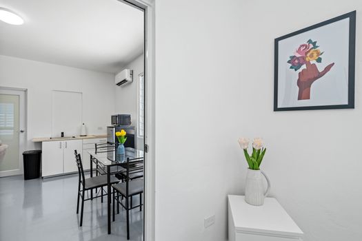 A sleek and airy dining area with a glass table, black chairs with slatted backs, and a vase of yellow flowers, featuring artworks of flowers, buildings, and abstract shapes on the walls.