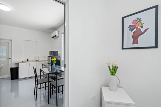 A sleek and airy dining area with a glass table, black chairs with slatted backs, and a vase of yellow flowers, featuring artworks of flowers, buildings, and abstract shapes on the walls.