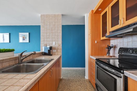 Fully equipped kitchen with double sink, tiled countertops, wooden cabinetry, and modern appliances including a stove and coffee maker.