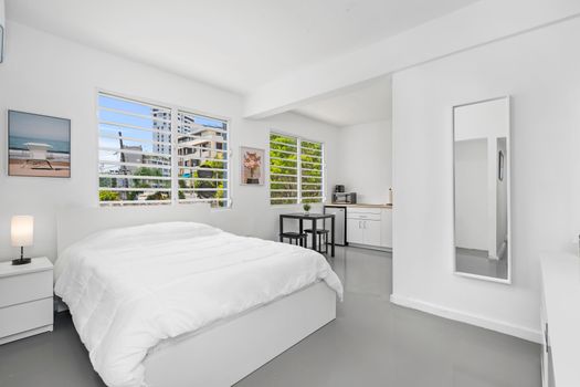 This cozy bedroom features large windows that offer stunning views of the cityscape. The room is immaculately clean and modern with white walls with large mirror and a neatly made white bed. A piece of modern artwork hangs on the blue wall adding an artistic touch to the room. A large window allows ample natural light to flood into space, enhancing its brightness. Outside the window are views of buildings indicating an urban setting.