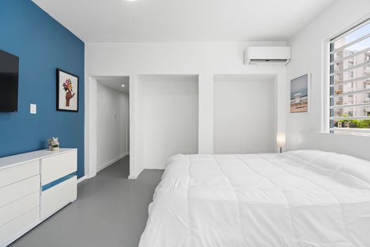 This modern minimalist room features sleek design and natural light. The blue accent wall adds a pop of color to the space. A piece of modern artwork hangs on the blue wall adding an artistic touch to the room. A large window allows ample natural light to flood into space, enhancing its brightness. Outside the window are views of buildings indicating an urban setting.