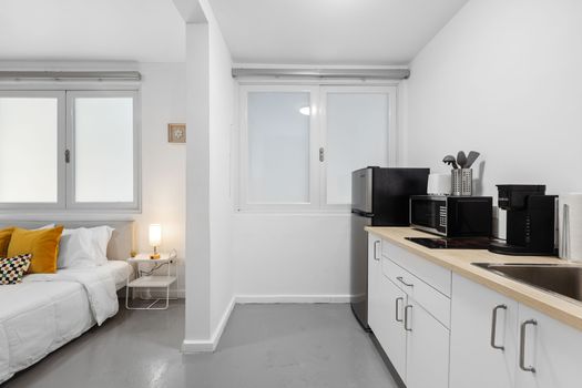 Cocina pequeña compacta adyacente al área de dormitorio con electrodomésticos modernos y gabinetes blancos.