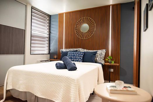 Discover comfort and style in this inviting bedroom. Featuring a warm wooden headboard with ambient lighting, a plush queen bed, and thoughtful blue accents, it's your perfect restful retreat.