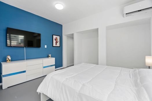 On the right side of bed there’s an entertainment unit featuring drawers and a mounted flat-screen TV against blue accent wall. The floor appears to be grey which complements both white walls & blue accent wall creating contrast yet harmonious look.