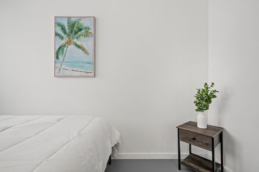 Relax in this cozy and bright bedroom by the sea, featuring a comfortable bed, a serene beach painting, and fresh greenery.