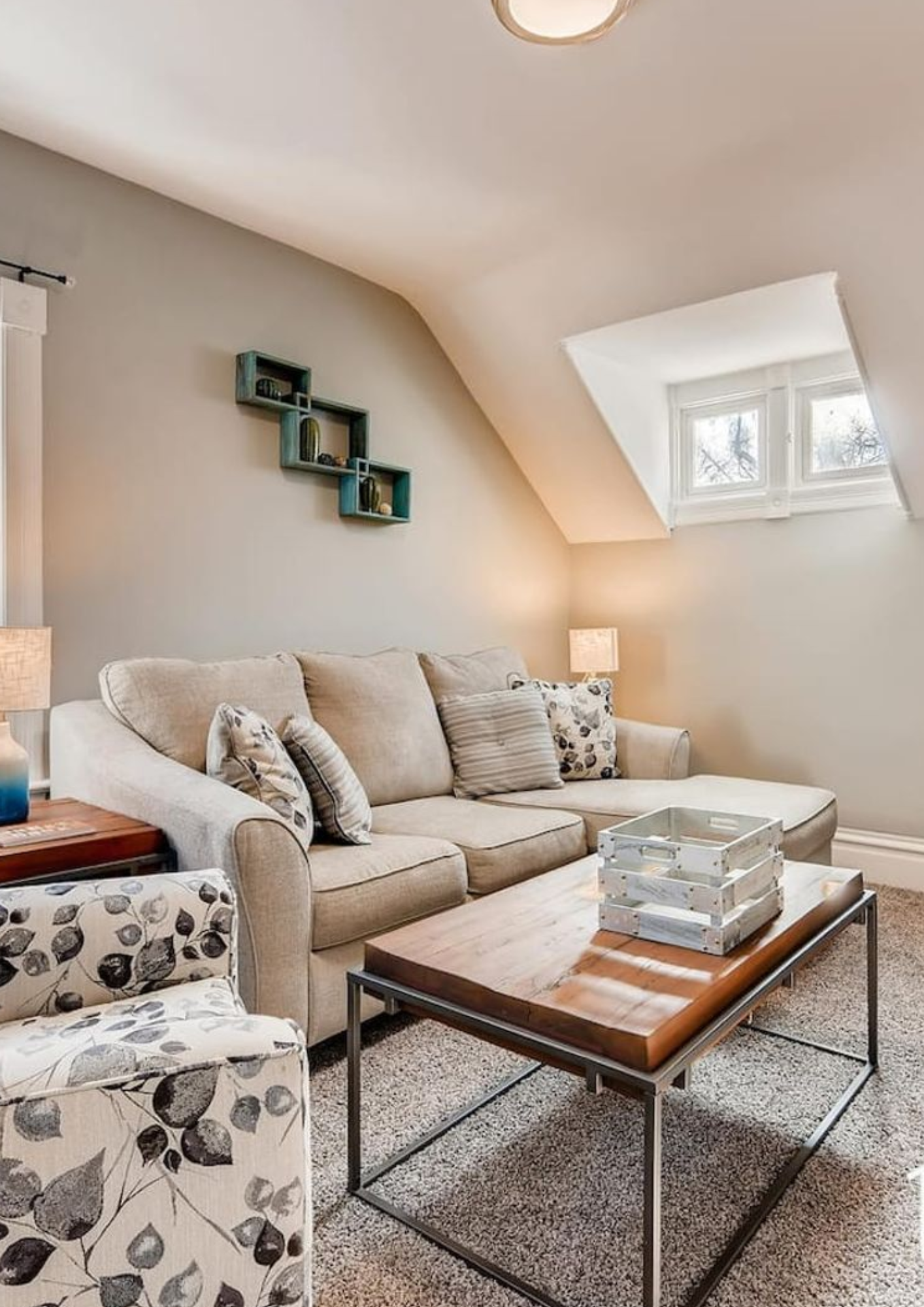 A well-lit living area featuring a beige l-shaped sofa with patterned pillows, a rustic wood coffee table on a grey carpet, and a sloped wall with a dormer window.