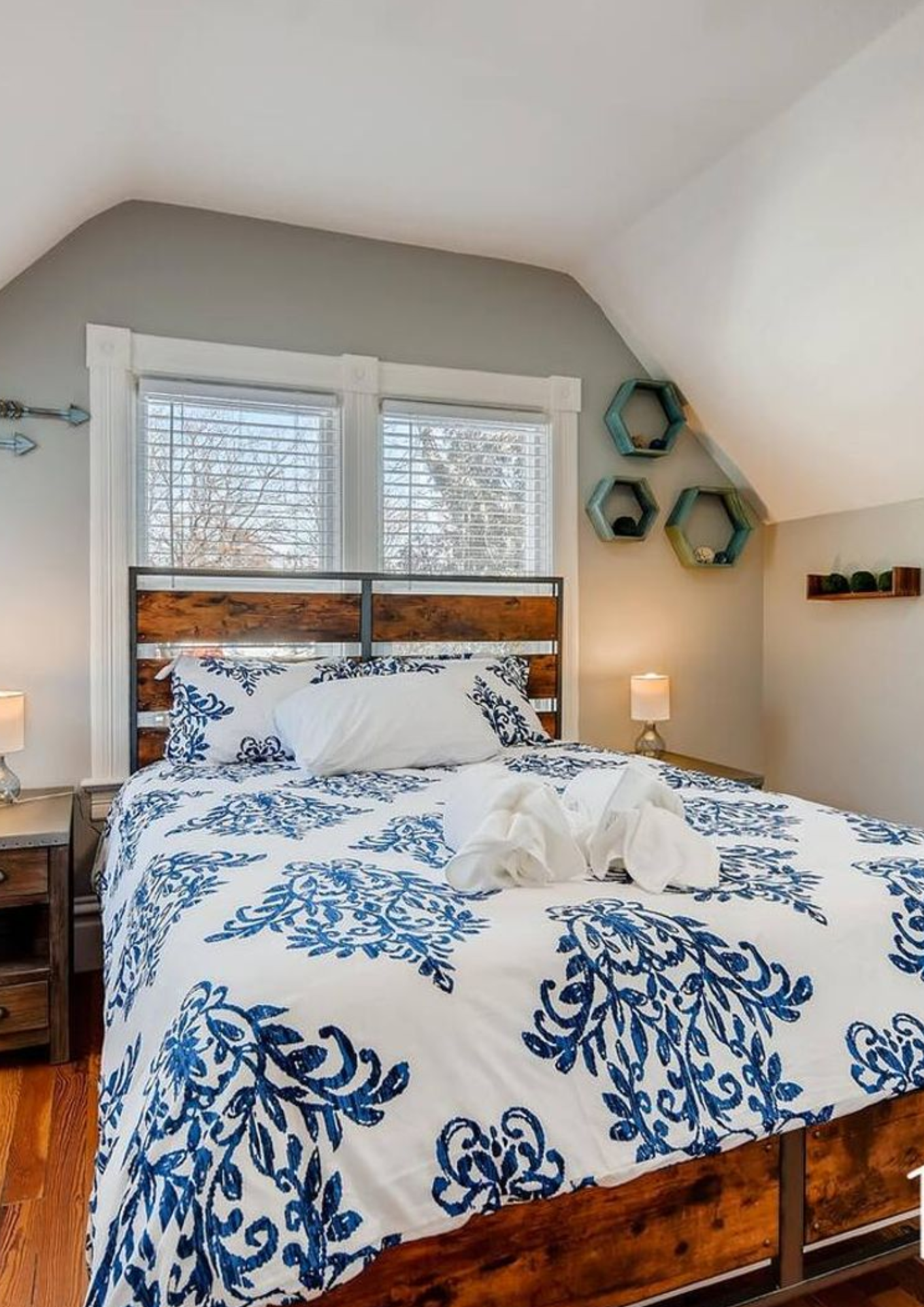 A bright bedroom with a queen bed, blue floral patterned duvet, and a reclaimed wood headboard flanked by industrial-style nightstands and hexagon wall shelves.
