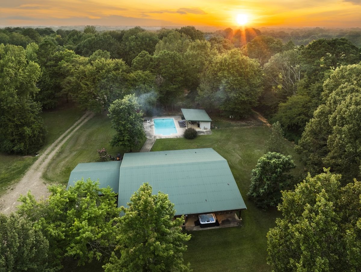 Aerial view of a cozy cabin surrounded by lush greenery at sunset, featuring a swimming