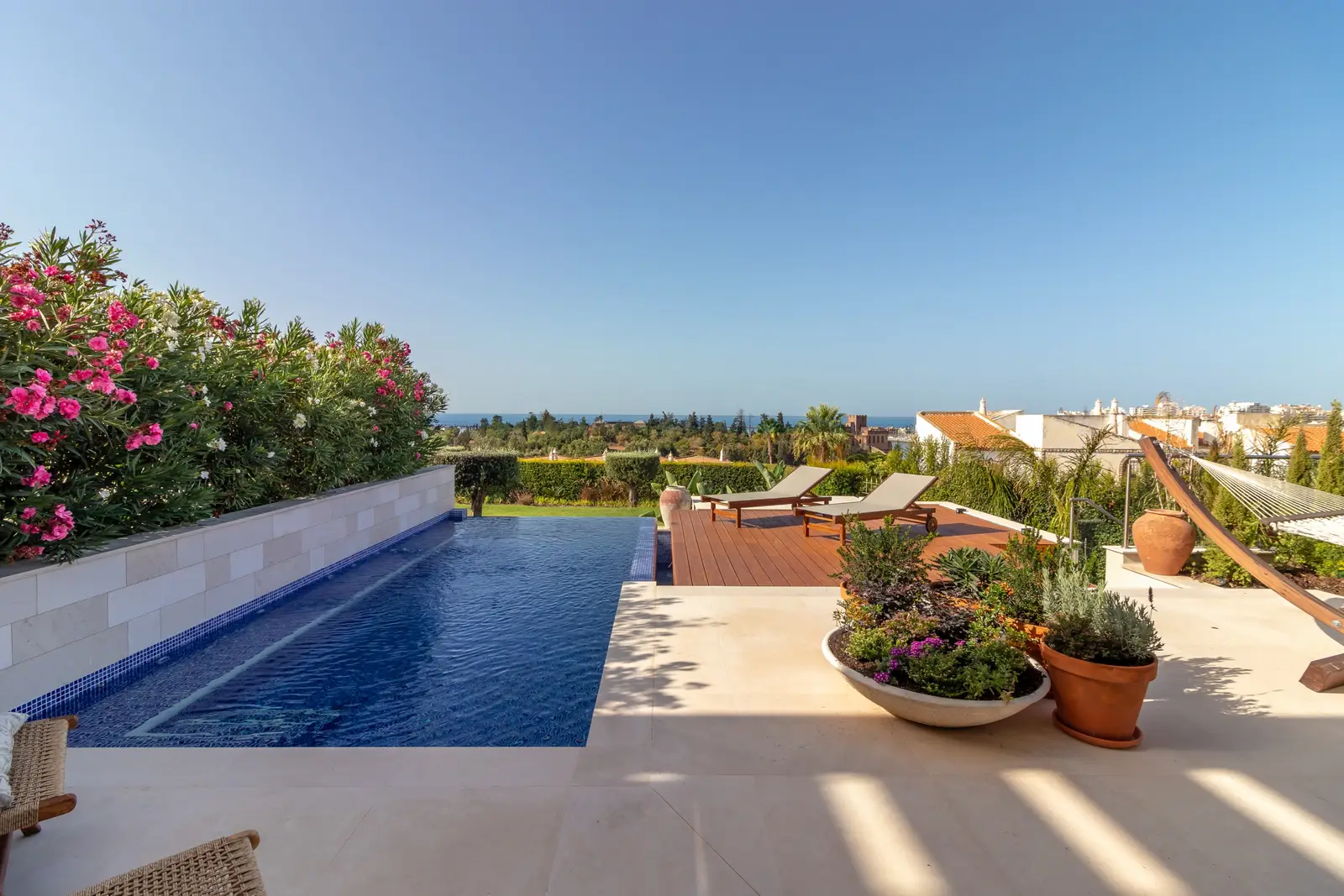 Blue-tiled infinity pool beside a stone terrace with planters, sun loungers and the sea beyond