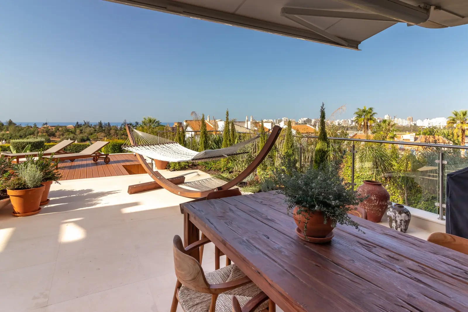 Wooden terrace table with potted olive, hammock and sea view beyond the glass balustrade