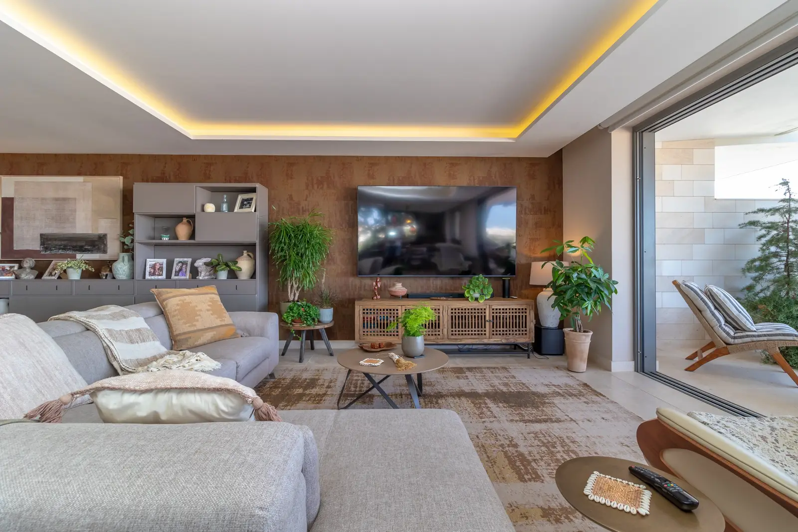 Grey corner sofa facing a wall-mounted television on a cork-clad wall, with plants and rugs