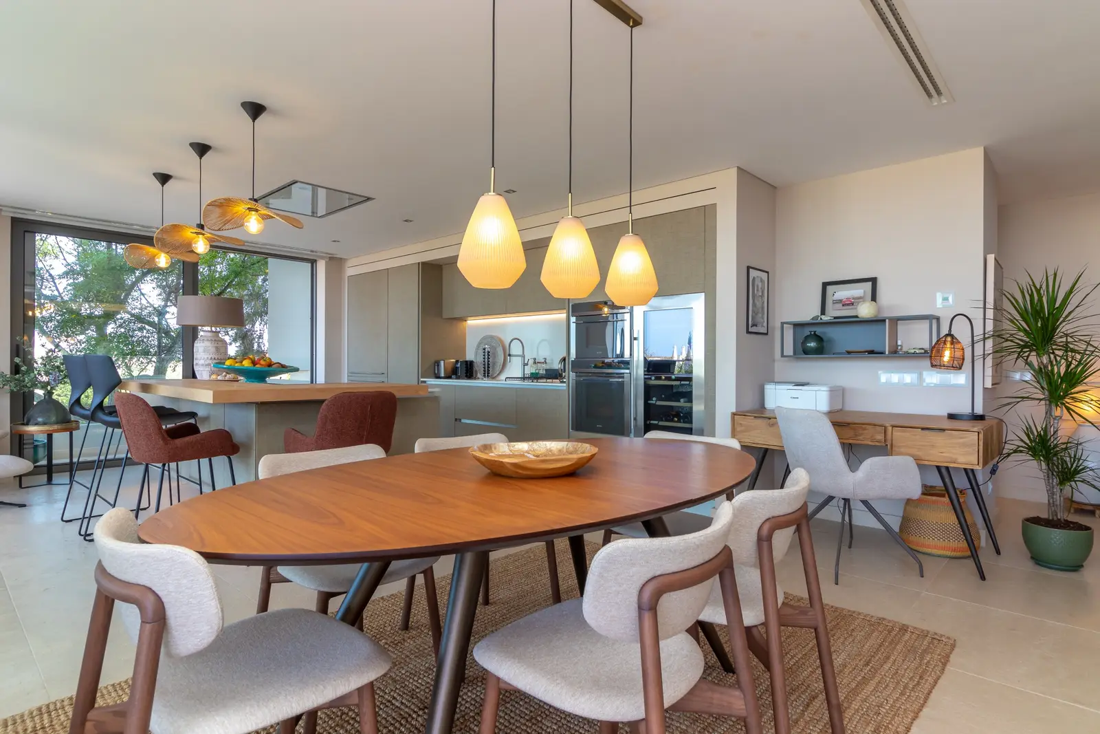 Oval wooden dining table with upholstered chairs, pendant lights and kitchen beyond
