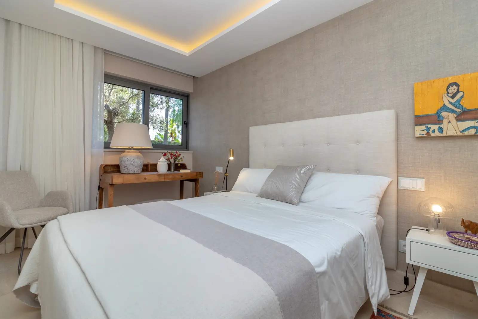 Bedroom with upholstered headboard, white bedding, armchair and window onto trees