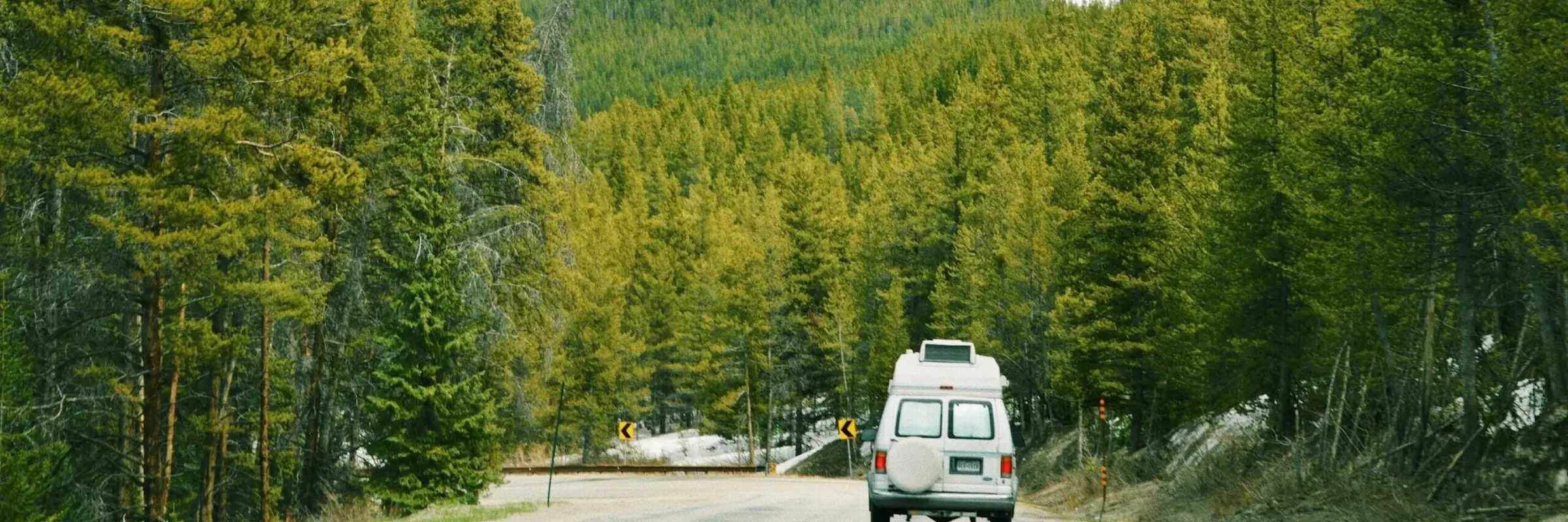 Camper van driving along a winding mountain road surrounded by dense evergreen forest and snow-covered slopes.