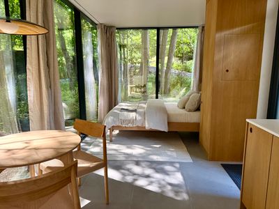 Open-plan guest studio: bed, dining, and forest views under one glass-wrapped roof.