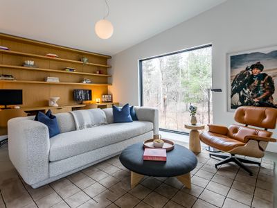 A bright, book-lined living area with a built-in oak shelving nook, plush sofa, and a leather lounge chair framing the forest through floor-to-ceiling glass.