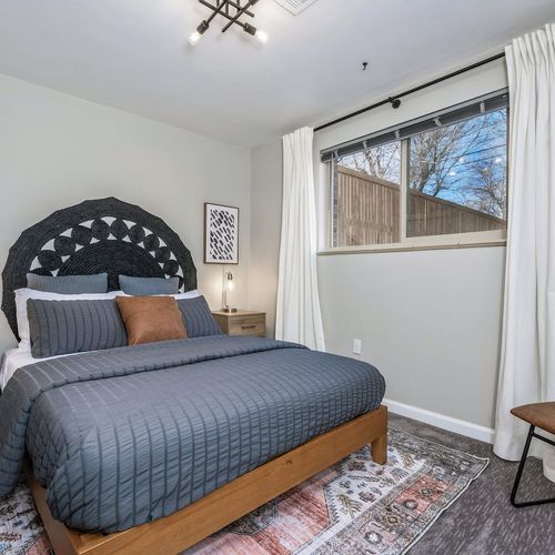 - queen bed with dark gray bedding pops against white curtains and wood accents
- large window lets in light while offering backyard views and privacy
- soft rug adds color comfort and personality to this serene sleep space