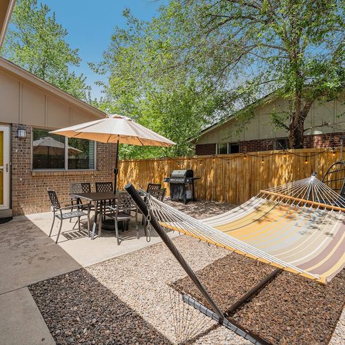 - view of the fenced in patio with hammock umbrella table and bbq setup
- sunshine filters through trees onto gravel paths and concrete seating zone
- enjoy summer days with breezy naps or meals just steps from the yellow door