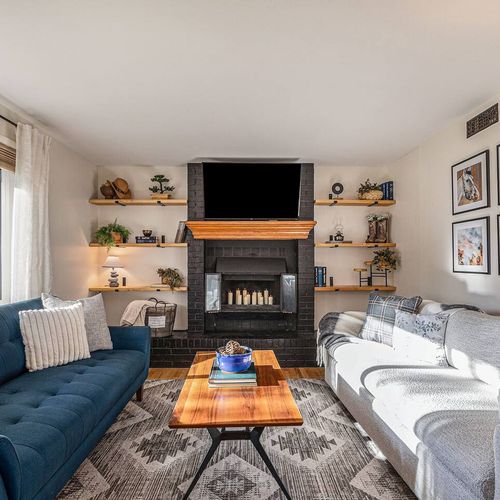 - cozy up by the black fireplace surrounded by soft couches and rustic wooden shelving
- neutral decor, patterned rug, and large tv create a perfect family space
- bright windows keep the space feeling open and airy all day