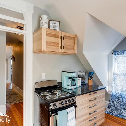- corner kitchenette includes a compact electric stove, prep space, and full utensil set
- natural light from the window enhances the warmth of the soft wood cabinetry
- coffee maker and toaster sit ready for quick breakfasts or late night snacks