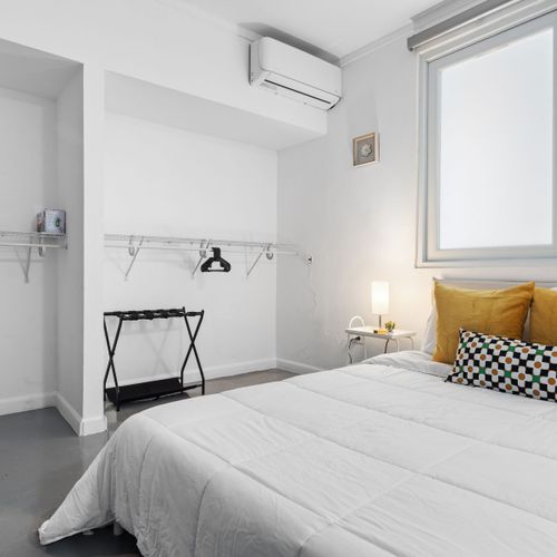 Dormitorio elegante y minimalista con un toque de color, que cuenta con una cómoda cama con almohadas estampadas en color mostaza, complementada con un práctico armario abierto y una moderna unidad de aire acondicionado para una comodidad óptima.