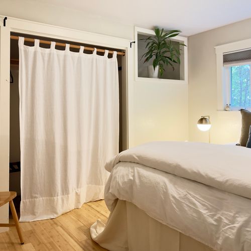 A cozy bedroom with a neatly made bed dressed in white bedding, a bedside lamp, a wooden chair, a curtained closet opening, light wood floors, and a small built-in shelf holding a potted plant.
