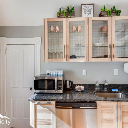 - mini kitchen setup with a microwave, coffee station, sink, and under counter dishwasher
- glass front cabinets hold essentials and add visual openness above black granite
- decorative greenery and clean design elevate this compact kitchen zone
