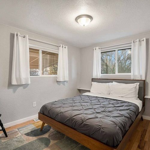 - queen bedroom with dual windows and dark gray comforter for a moody yet cozy feel
- modern decor paired with light wood flooring and white blackout curtains throughout
- corner chair and stylish bed frame offer both function and flair