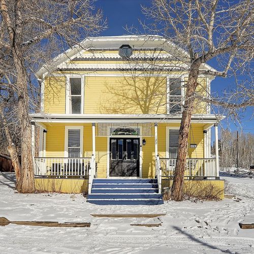 Charming yellow victorian style home with black front door, wraparound porch, and snowy winter curb appeal.
