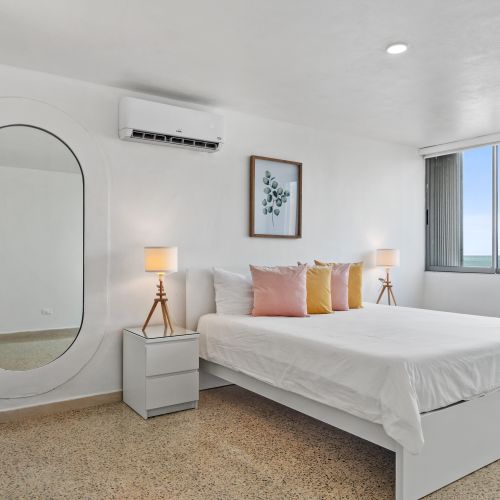 Bedroom with queen bed, white bedding, nightstand, modern lamp, and large oval mirror. Air conditioning unit visible.