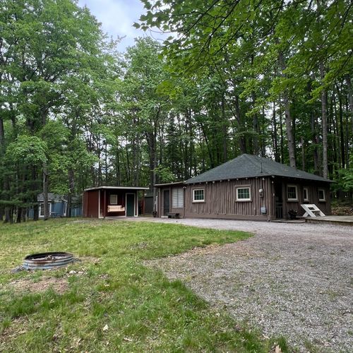 EXTERIOR:  A broader view of the front of the cabin, driveway, and bonfire ring in the summer.