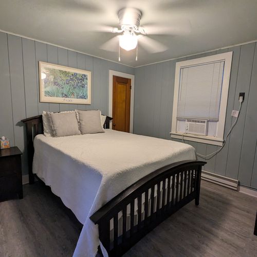 BEDROOM #5 (BEACH HOUSE, MAIN FLOOR):  Private bedroom with a queen-size bed, window A/C unit, ceiling fan, dresser, nightstand, lamp, mirror, and a travel crib.