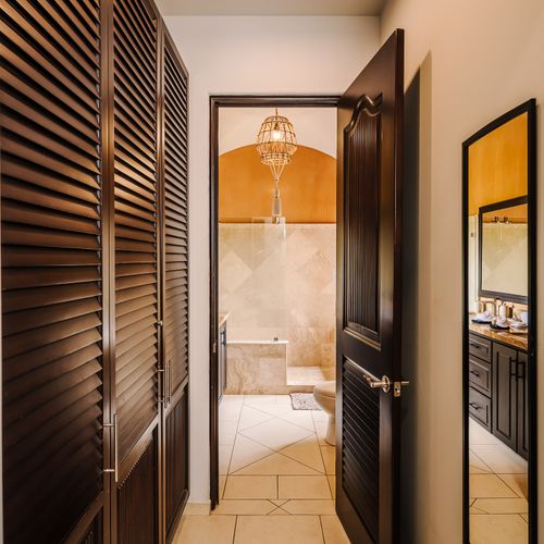 Walk-through dressing alcove with louvered closets, rattan pendant light, and view into ochre travertine master bath.