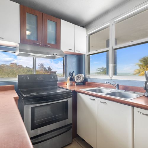 Experimente una cocina moderna con accesorios elegantes y abundante luz natural, que cuenta con fregadero, estufa y horno blancos, y una vista pintoresca de la playa a través de la ventana.