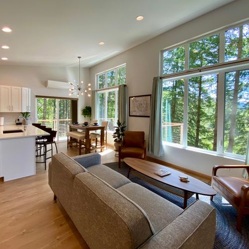 The whole picture — open-plan living, dining, and kitchen wrapped in floor-to-ceiling forest views on every side