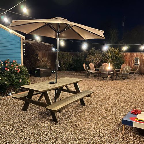 - magical evening ambiance with glowing string lights and a cozy fire pit gathering area
- picnic table and umbrella provide a great space for nighttime snacks or games
- vibrant shed, blooming flowers, and cornhole boards add charm and fun to the sc