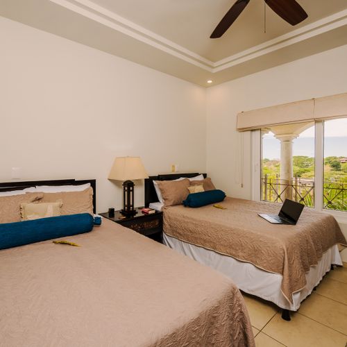 Second bedroom with two beige-quilted beds, turquoise bolsters, and arched window view to palms and ocean glimpse.