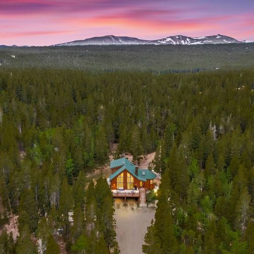 Aerial view of a secluded mountain cabin surrounded by forest, set against a stunning sunset and alpine backdrop.
