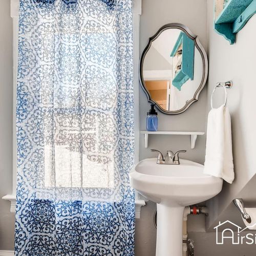 - patterned blue curtain brings brightness to this bathroom with a classic pedestal sink
- oval mirror, white shelving, and towel hook keep essentials within easy reach
- light gray walls and teal accents give the space a fresh and airy feel