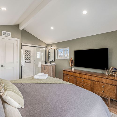 Step directly into a modern ensuite bathroom with glass shower & sleek vanity - clean lines, vaulted ceiling, and soft lighting create an inviting, restful bedroom escape - enjoy a smart tv, dresser storage, and curated accents all in one spot