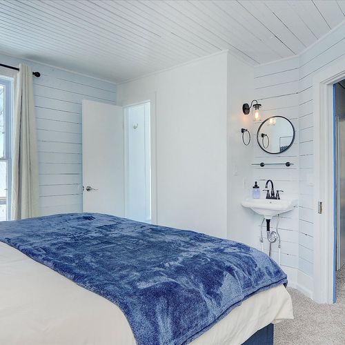 - bright bedroom with a cozy bed, soft blue accents, and a wall mounted sink vanity
- large window lets in plenty of natural light and frames charming outdoor views
- clean lines, wood paneled walls, and modern fixtures offer a peaceful retreat