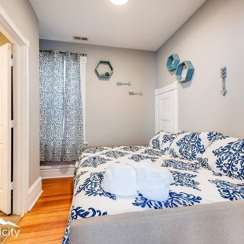 - cozy bedroom with a queen bed dressed in blue botanical bedding and soft lighting
- unique wall decor and hexagon shelves bring playful personality to the space
- hardwood floors and a private curtain lined window complete the look