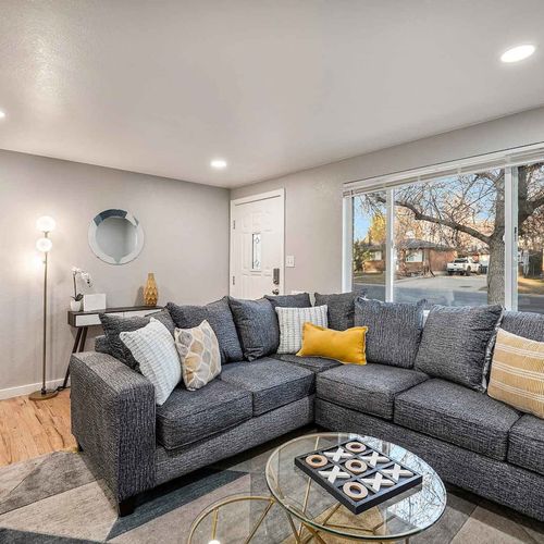 - cozy living room with oversized sectional, bright windows, and warm wood flooring
- pops of yellow and a round mirror bring a modern touch to the neutral palette
- comfortable space for relaxing, reading, or catching your favorite shows