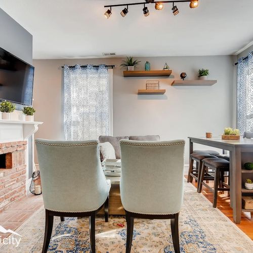 - cozy living room with a brick fireplace & soft chairs facing a sleek mounted tv setup
- dining table with stools sits near bright windows for light filled meals or games
- floating wall shelves and track lighting enhance the warm inviting ambiance